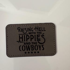 Puede incluir: Un parche rectangular de cuero gris con la frase "Raising Hell with the Hippies and the Cowboys" en letras negras. Cinco pequeñas estrellas en la parte inferior. El parche tiene un borde cosido.