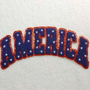 Puede incluir: Un parche azul y rojo con la palabra "AMERICA" en un diseño audaz y arqueado. Las letras son azules con estrellas blancas, contorneadas en purpurina roja. El parche es probablemente para ropa o accesorios.