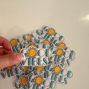 May include: White embroidered patches with the words "GOOD VIBES" in teal letters, a yellow sun graphic, and a black outline. The patches are rectangular and have a white background. The image shows a hand holding one patch, with many more patches in the background.