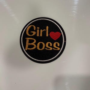 Pode incluir: Patch bordado circular com as palavras "Girl Boss" em letras douradas, um coração vermelho e um fundo preto. O patch tem uma borda branca.