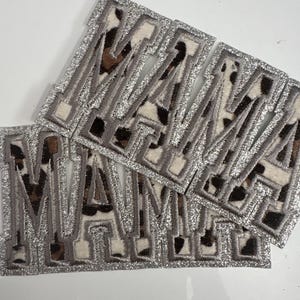 May include: Embroidered patches spelling "MAMA" in a stacked arrangement. Each patch features a silver glitter border, a gray outline, and a leopard print pattern in brown, black, and cream colors. These patches are ideal for DIY projects.