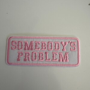 May include: A rectangular patch with the phrase "SOMEBODY'S PROBLEM" embroidered in pink on a white background. The patch has a pink border and is likely designed to be sewn onto clothing or accessories.