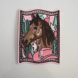 May include: Embroidered patch featuring a brown horse head, a pink cowboy hat, and cacti. The patch has a pink border with a black stitched frame. White stars are scattered across the pink background. The patch is rectangular with a wavy top edge.