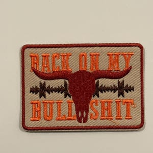 Könnte beinhalten: Gestickter Aufnäher mit beigem Hintergrund, rotem Bullenschädel und dem Text "BACK ON MY BULL SHIT" in Orange. Der Aufnäher hat einen braunen Rand und ein Stacheldrahtdesign.