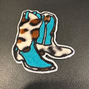 May include: An embroidered patch featuring a pair of cowboy boots. The boots have a turquoise shaft and a brown and white animal print design. The patch is outlined with a white border.