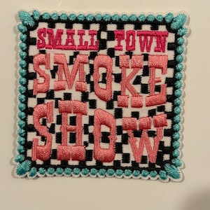 Peut inclure: Écusson brodé carré avec l'inscription rose "SMALL TOWN SMOKE SHOW". Le patch a un fond à carreaux noir et blanc, une bordure turquoise et un dos blanc. Le patch est de forme carrée.