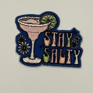 May include: Embroidered patch featuring a pink margarita cocktail with a lime wedge, rimmed with salt. The patch is blue with the words "STAY SALTY" in a stylized font. Decorative floral elements are included.
