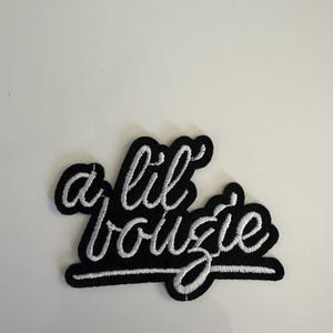 May include: A black embroidered patch with the phrase "a lil bougie" in white cursive lettering. The patch has a black border and is likely designed to be ironed on or sewn onto clothing or accessories.