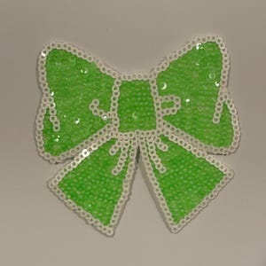 May include: A vibrant green sequined bow with a white beaded trim. This decorative bow is likely for embellishing clothing or craft projects. The sequins provide a shiny, textured finish to the bow.