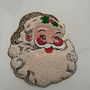 May include: A Santa Claus face-shaped Christmas decoration. The face is light pink with red cheeks and a red mouth. The beard and hat are off-white with a gold glitter trim. The hat has a leopard print pattern and holly berries.
