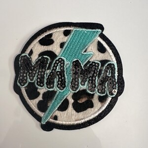 Puede incluir: Un parche redondo con la palabra "MAMA" en lentejuelas negras, un rayo turquesa y un fondo de estampado de vaca en blanco y negro. El parche tiene un borde negro.