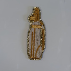 May include: Embroidered patch in the shape of a golf bag. The patch has a gold glitter outline and accents, with a beige and gold colour scheme. The design includes vertical lines and a horizontal band.