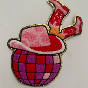 Op de afbeelding: Een kleurrijke patch met een discobal met een cowboyhoed en laarzen. De hoed is roze met een wit patroon, de laarzen zijn rood en de discobal is rood en paars. De patch is goudkleurig omlijnd.