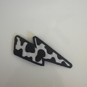 May include: Black and white lightning bolt patch with a cow print design. The patch is a fabric applique with a black border and a white and black pattern resembling a cow's spots. The lightning bolt shape is a popular design element.