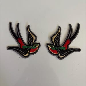 Puede incluir: Dos parches bordados con golondrinas en vuelo. Las aves tienen alas negras, pechos rojos y detalles verdes, con detalles dorados. Los parches están delineados en gris.