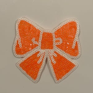May include: An orange sequined bow with a white beaded border. The bow is a decorative item, possibly for crafts or embellishment. The sequins are closely packed, giving a textured appearance.