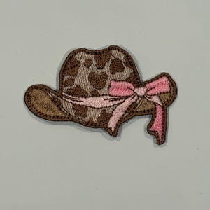 May include: Embroidered patch of a brown cowboy hat with a pink bow. The hat features a tan and brown spotted pattern. The patch is designed to be ironed or sewn onto fabric, suitable for decorating clothing or accessories.