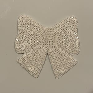 May include: A white sequined bow, likely for embellishment, is centered against a neutral background. The bow's shape is clearly defined, with each sequin reflecting light. The design suggests a craft or fashion accessory.