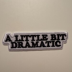 Puede incluir: Un parche rectangular con la frase "A LITTLE BIT DRAMATIC" bordada en hilo negro. El parche tiene un borde blanco y está diseñado para ser cosido o planchado en ropa o accesorios.