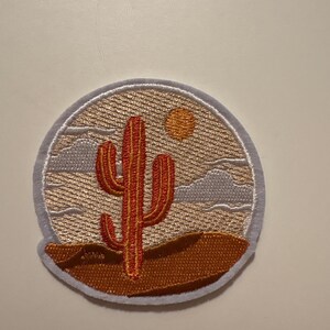 Op de afbeelding: Ronde geborduurde patch met een woestijnlandschap. Het heeft een rode cactus, een gele zon en bruin zand. De patch heeft een witte rand en is ontworpen voor toepassing op stoffen items.
