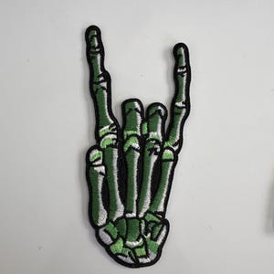 Może przedstawiać: Haftowana naszywka przedstawiająca zieloną dłoń szkieletu w geście 'rock on'. Naszywka ma czarną obwódkę i szczegółowe przeszycia. Idealna na kurtki, torby lub inne przedmioty z tkaniny. Białe tło.
