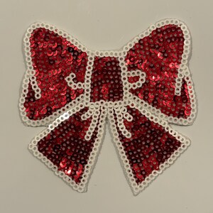May include: A decorative bow made of red sequins with a white beaded border. The bow is a classic shape, with two loops and two streamers. The sequins catch the light, creating a sparkling effect.