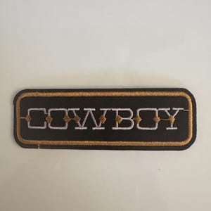 May include: Embroidered patch with the word "COWBOY" in white, outlined in gold, on a black rectangular background. The patch has a gold border and is likely designed to be ironed or sewn onto clothing or accessories.