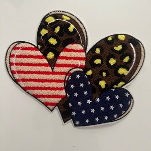 May include: A cluster of heart-shaped patches. One patch displays red and white stripes, another has a blue background with white stars, and two have a brown leopard print with yellow spots. Each patch has a white stitched border.