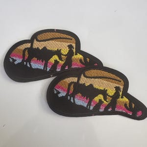 May include: Two embroidered patches in the shape of a cowboy hat. The patches feature a silhouette of a cowboy, horse, and cactus against a sunset background with pink, yellow, and blue hues. The hat brim is brown, and the patches have a black border.
