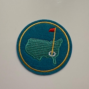 May include: A round, teal patch with a gold border. Inside, a green embroidered outline of the United States with a golf pin and red flag. The patch is likely for apparel or accessories, representing a golf theme.