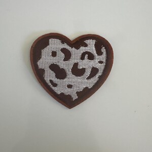 May include: A heart-shaped patch with a brown border and a white and brown leopard print design. The patch is likely made of fabric and could be used to decorate clothing or accessories.