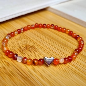 Puede incluir: Un brazalete de piedra de cornalina roja con un dije de corazón plateado. El brazalete está hecho de pequeñas cuentas redondas y está enhebrado en un cordón elástico transparente.