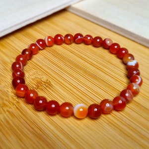 Könnte beinhalten: Ein rotes Karneol-Armband mit weißen Streifen auf den Perlen. Das Armband besteht aus natürlichen Steinen und liegt auf einer Holzoberfläche.