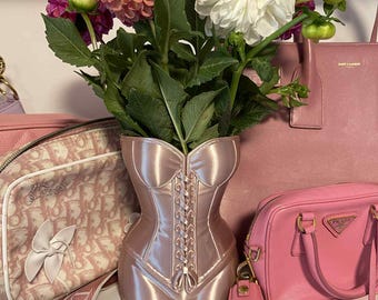 Peut inclure: Un vase rose en forme de corset contient un bouquet de dahlias roses et blancs. Le vase a une finition satinée et un design à lacets. D'autres sacs à main roses sont en arrière-plan.