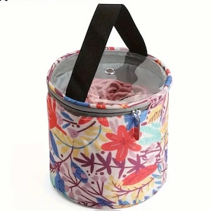 May include: A circular knitting bag with a floral pattern, black handle, and zip closure. The bag has a clear top, revealing the contents. The design features red, blue, yellow, and purple flowers and foliage.