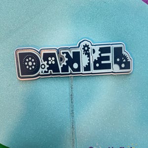 May include: A blue and silver cake topper with the name "DANIEL" spelled out in black letters with gears and other mechanical elements. The topper is on a silver glitter stick.