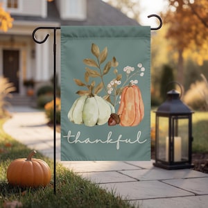 Puede incluir: Una bandera de jardín rectangular con fondo verde salvia y una ilustración en acuarela de una calabaza verde pálido, una calabaza naranja, una bellota y follaje marrón. La palabra thankful está impresa en letra cursiva blanca.