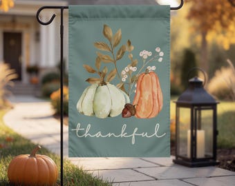 Könnte beinhalten: Eine rechteckige Gartenflagge mit salbeigrünem Hintergrund und einer Aquarellillustration eines hellgrünen Kürbisses, eines orangefarbenen Kürbisses, einer Eichel und braunem Laub. Das Wort thankful ist in weißer Schreibschrift aufgedruckt.