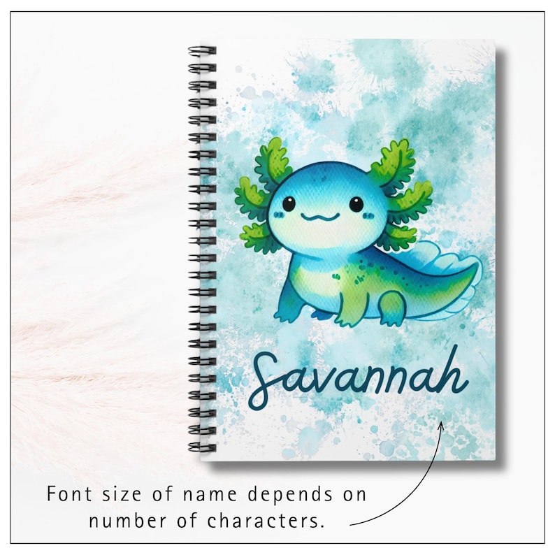 Image of notebook cover.  A cheerful cartoon axolotl with round, smiling face and rosy cheeks. Its body is light teal with darker green spots and shading, and it has large frilly gills on each side of its head in shades of green and teal. Background is a soft watercolor-style wash of teals and green, giving a dreamy, splashy effect. At the bottom, the name “Savannah” is written in playful, dark blue script lettering. Text says, with an arrow, "Font size of name depends of number of characters."