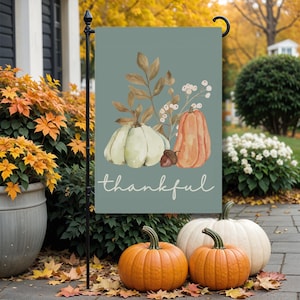 May include: A teal garden flag with a watercolor design of pumpkins, leaves, and the word "thankful." The flag is displayed outdoors with pumpkins and fall foliage.