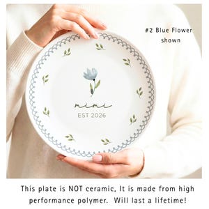 May include: White plate with a blue floral design and the word "mimi" with "EST 2026" underneath. The plate has a scalloped edge with small green leaf accents. The text "#2 Blue Flower shown" is also visible.