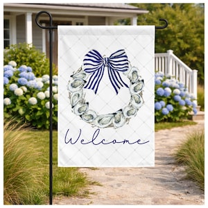 May include: A white garden flag featuring a watercolor oyster shell wreath with a navy blue and white striped bow. The word "Welcome" is written in navy blue script below the wreath. The flag is displayed on a black metal stand.