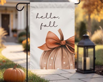 May include: A white garden flag featuring the text hello, fall in black script lettering above a watercolor illustration of a light brown pumpkin with white polka dots and a large, tied orange bow on its stem, hanging from a black metal stand.