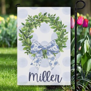 May include: A garden flag featuring a green leafy wreath with a blue floral bow on a white background with blue polka dots. The name "Miller" is printed in navy blue script at the bottom. The flag is displayed on a black metal stand.