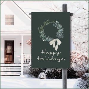 Op de afbeelding: Een donkergroene tuinvaandel met een aquarelkrans en een wit strikdesign. De woorden "Happy Holidays" zijn in een cursief lettertype geschreven. De vlag wordt getoond voor een wit huis in een besneeuwde omgeving.