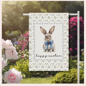 Peut inclure: Un drapeau de jardin avec une illustration à l'aquarelle d'un lapin portant un nœud papillon bleu. Le drapeau a un fond blanc avec un motif de feuilles vertes et les mots "happy easter". Le drapeau est affiché sur un support en métal blanc.