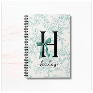 Op de afbeelding: Spiraalgebonden notitieboek met een bloemmotief in lichtgroen en wit. De letter "H" is gecentreerd, versierd met een teal strik. De naam "haley" staat eronder in een scriptlettertype. Zwarte spiraalbinding.
