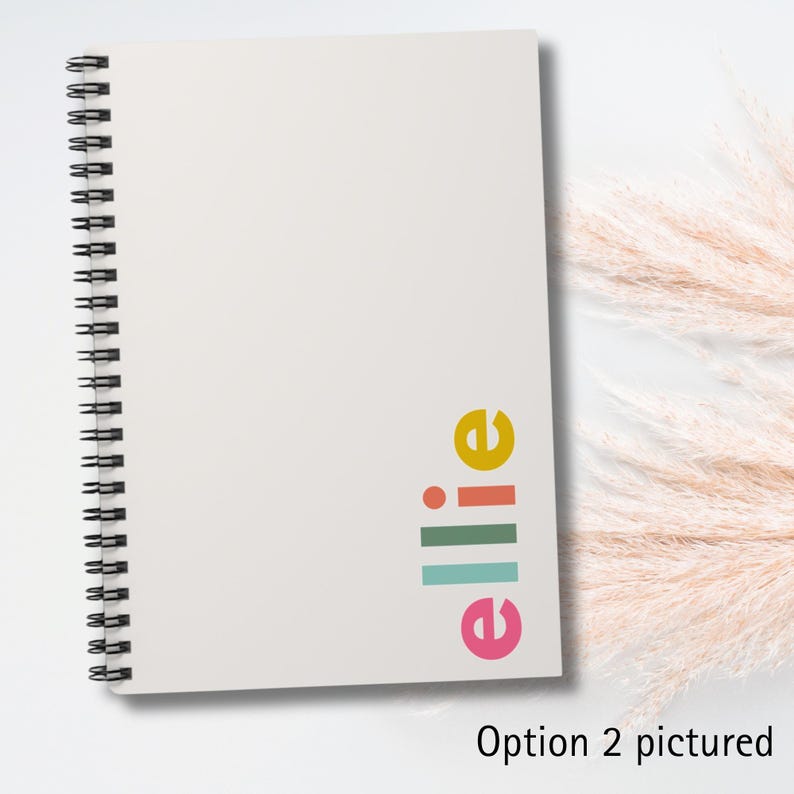 A white spiral bound notebook with a colorful logo on the front. The logo is a stylized word 'elle' with a yellow 'e', a pink 'e', a green line, a blue line, a red dot, and a green line. The notebook is on a white surface with a white background. Option 2 pictured.