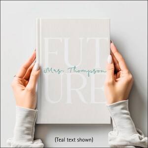 A woman's hands are shown holding a light beige hardcover journal. The journal cover has the large word “FUTURE” printed in a soft white font across the front. The letters F, U, and T are on the top row with the letters U, R, and E below. Across the middle, the name “Mrs. Thompson” is written in a teal cursive script. The person holding the journal is wearing a gray sweatshirt, and the background is softly blurred. Text says: (Teal font shown).