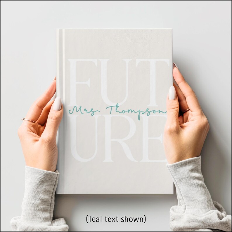 A woman's hands are shown holding a light beige hardcover journal. The journal cover has the large word “FUTURE” printed in a soft white font across the front. The letters F, U, and T are on the top row with the letters U, R, and E below. Across the middle, the name “Mrs. Thompson” is written in a teal cursive script. The person holding the journal is wearing a gray sweatshirt, and the background is softly blurred. Text says: (Teal font shown).